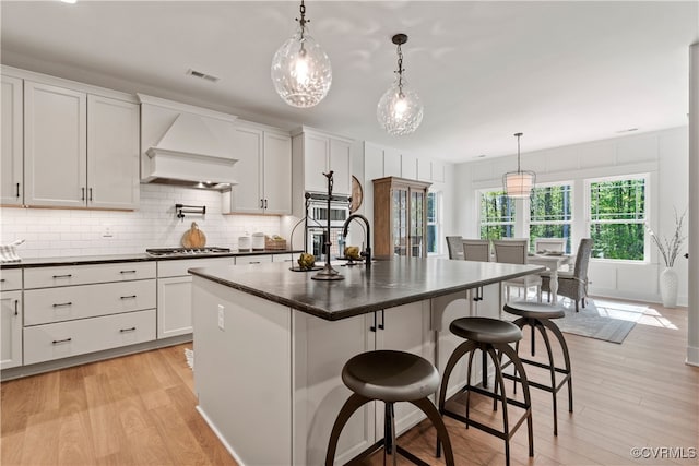 kitchen featuring custom range hood, light hardwood / wood-style floors, a kitchen island with sink, and white cabinetry