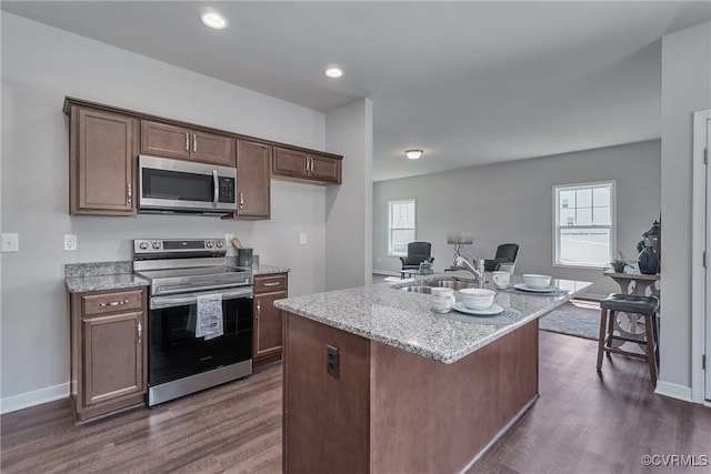 kitchen featuring light stone counters, dark wood-style floors, a wealth of natural light, appliances with stainless steel finishes, and a kitchen island with sink