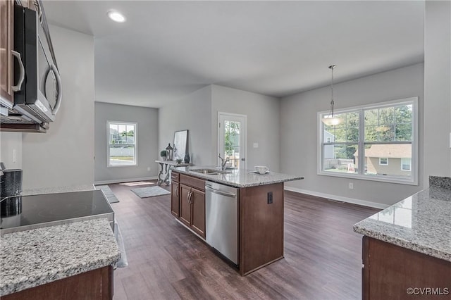 kitchen with pendant lighting, appliances with stainless steel finishes, dark wood-type flooring, a kitchen island with sink, and light stone countertops