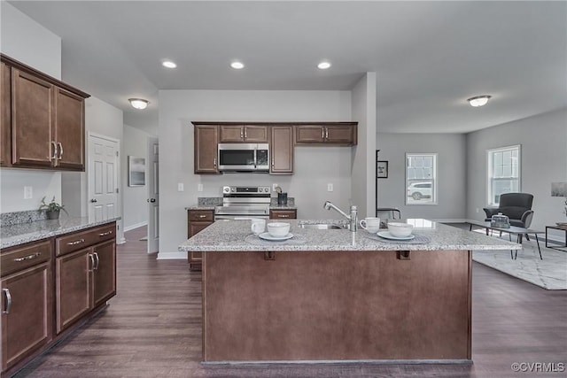 kitchen with stainless steel appliances, dark brown cabinets, a kitchen island with sink, and light stone countertops