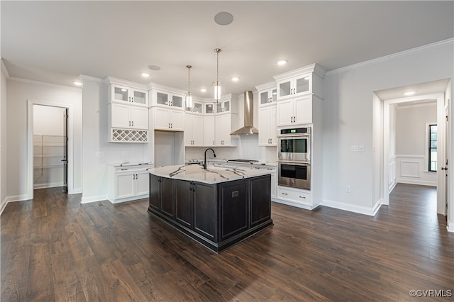 kitchen featuring an island with sink, decorative light fixtures, wall chimney exhaust hood, white cabinetry, and stainless steel appliances