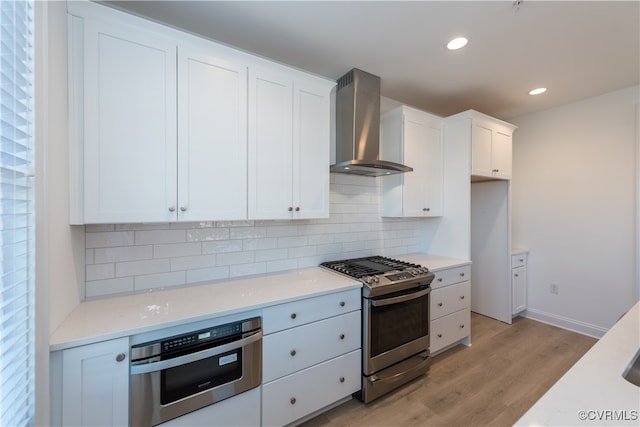 kitchen with wall chimney exhaust hood, decorative backsplash, light wood-type flooring, appliances with stainless steel finishes, and white cabinetry