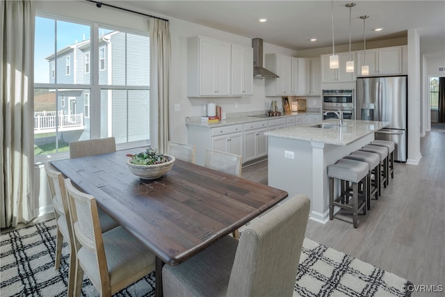 kitchen with white cabinets, an island with sink, decorative light fixtures, wall chimney exhaust hood, and stainless steel appliances