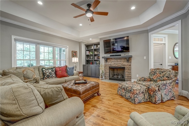 living room featuring light hardwood / wood-style floors, ceiling fan, a raised ceiling, a stone fireplace, and ornamental molding