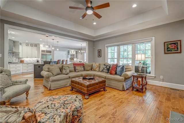 living room with ceiling fan with notable chandelier, light hardwood / wood-style floors, crown molding, and a raised ceiling