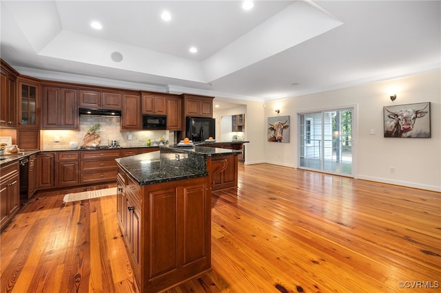 kitchen featuring light wood-type flooring, black appliances, a center island, a raised ceiling, and dark stone counters