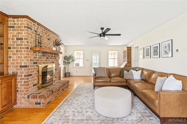 living room featuring a brick fireplace, a textured ceiling, light wood-type flooring, and ceiling fan