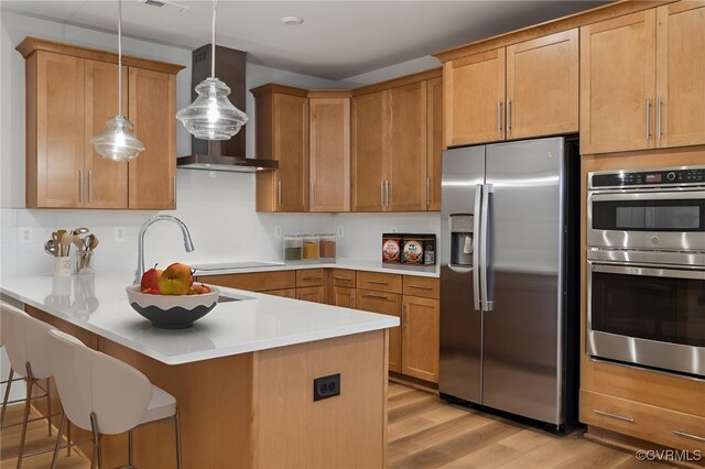 kitchen featuring wall chimney range hood, pendant lighting, light wood-type flooring, appliances with stainless steel finishes, and a kitchen breakfast bar