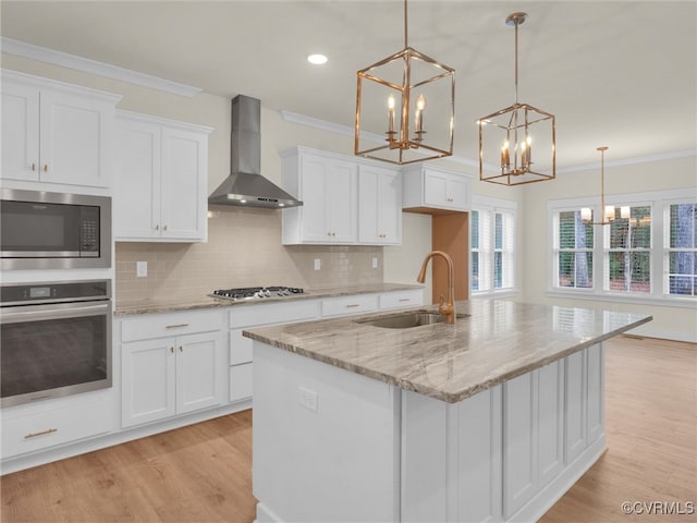 kitchen with appliances with stainless steel finishes, sink, wall chimney range hood, decorative light fixtures, and an island with sink