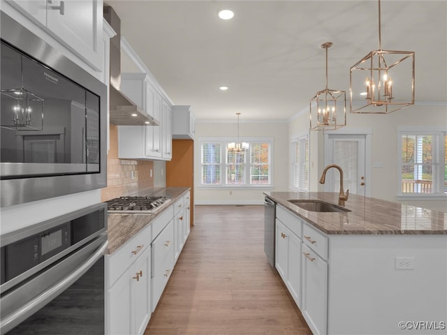 kitchen with a kitchen island with sink, white cabinets, wall chimney range hood, sink, and appliances with stainless steel finishes