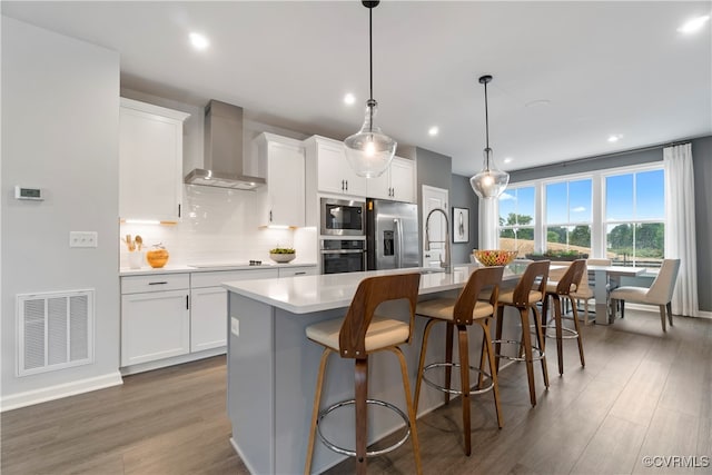 kitchen featuring wall chimney range hood, white cabinets, an island with sink, pendant lighting, and stainless steel appliances