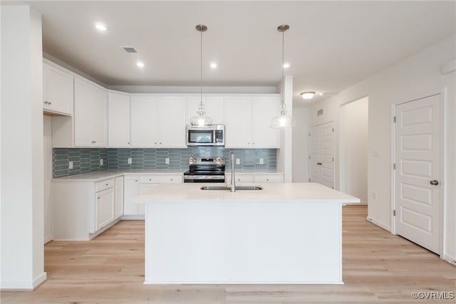 kitchen with appliances with stainless steel finishes, light wood-type flooring, decorative light fixtures, white cabinets, and a kitchen island with sink