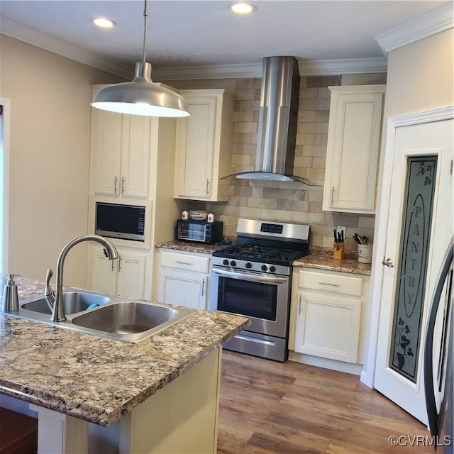 kitchen with light stone countertops, wall chimney range hood, appliances with stainless steel finishes, decorative light fixtures, and a center island with sink