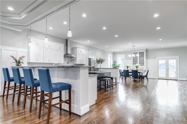 kitchen featuring stainless steel fridge, white cabinetry, crown molding, wall chimney exhaust hood, and decorative light fixtures