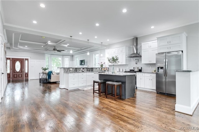 kitchen featuring wall chimney range hood, appliances with stainless steel finishes, white cabinetry, coffered ceiling, and a breakfast bar area