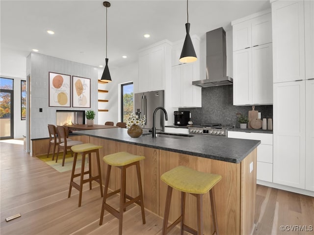 kitchen featuring wall chimney range hood, an island with sink, white cabinetry, light hardwood / wood-style floors, and sink