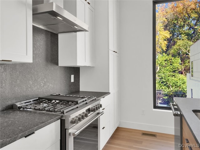 kitchen with tasteful backsplash, appliances with stainless steel finishes, white cabinetry, light wood-type flooring, and wall chimney exhaust hood