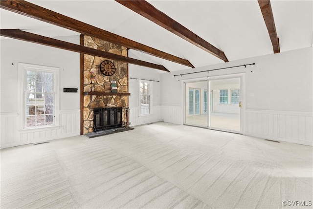 unfurnished living room featuring carpet, plenty of natural light, a wainscoted wall, a stone fireplace, and lofted ceiling with beams