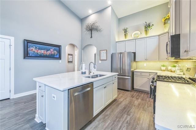 kitchen with a towering ceiling, stainless steel appliances, sink, an island with sink, and dark wood-type flooring