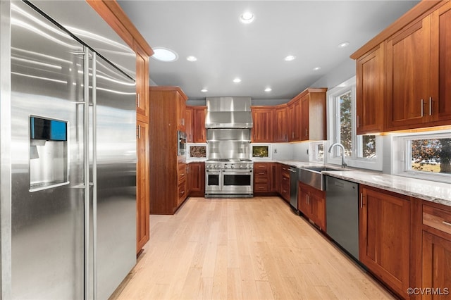 kitchen featuring light stone countertops, light wood-type flooring, high end appliances, wall chimney range hood, and sink