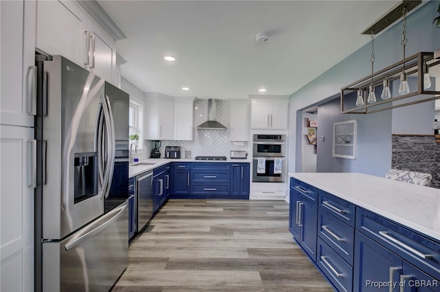 kitchen featuring white cabinets, wall chimney range hood, light hardwood / wood-style flooring, blue cabinetry, and stainless steel appliances