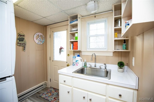 kitchen with a paneled ceiling, a baseboard heating unit, sink, stacked washer / drying machine, and white cabinetry