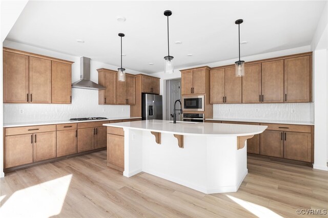 kitchen with a center island with sink, wall chimney range hood, hanging light fixtures, light wood-type flooring, and stainless steel appliances