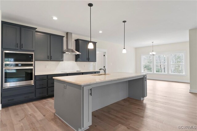 kitchen with a kitchen island with sink, wall chimney range hood, sink, hanging light fixtures, and light wood-type flooring