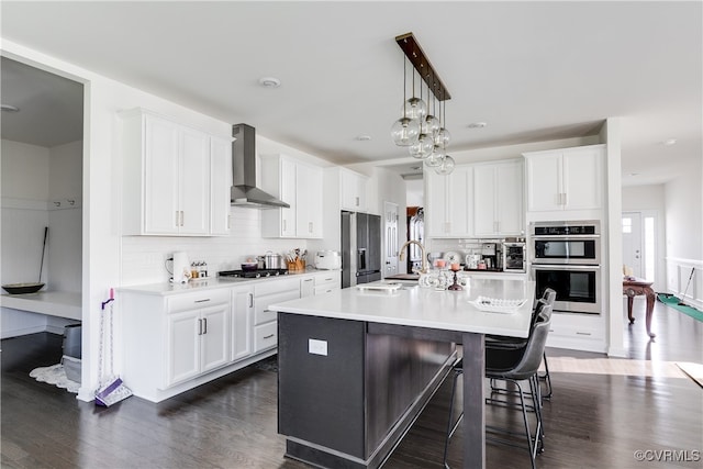 kitchen featuring white cabinetry, wall chimney range hood, an island with sink, pendant lighting, and appliances with stainless steel finishes