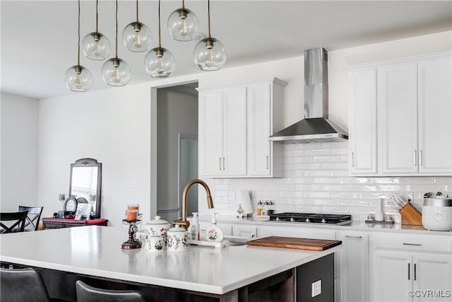 kitchen with a kitchen island with sink, a kitchen breakfast bar, hanging light fixtures, wall chimney exhaust hood, and white cabinetry