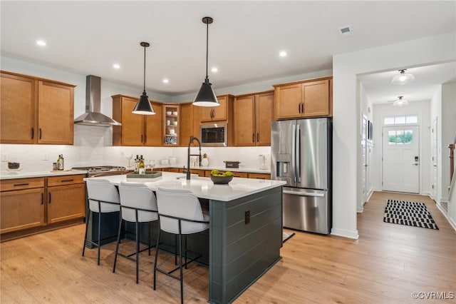 kitchen with brown cabinetry, light wood-style flooring, an island with sink, stainless steel appliances, and wall chimney range hood