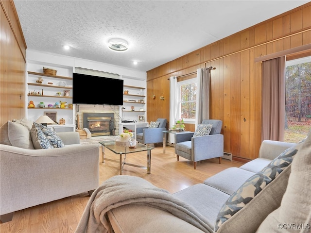 living room with a textured ceiling, built in features, light hardwood / wood-style floors, a stone fireplace, and wood walls