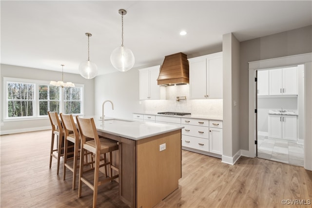 kitchen featuring premium range hood, sink, light wood-type flooring, an island with sink, and white cabinetry