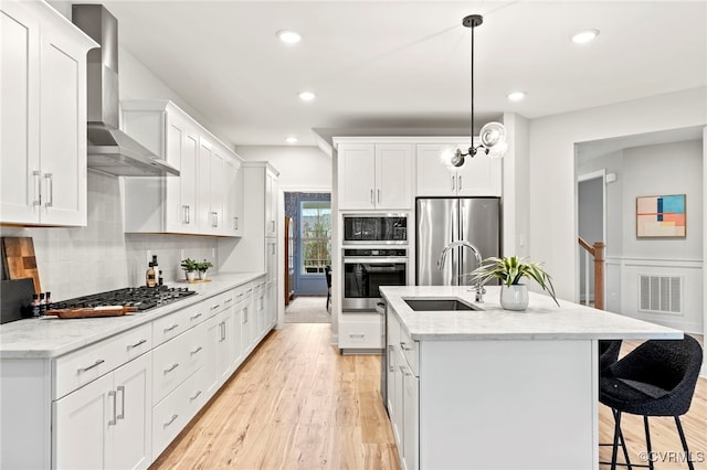 kitchen with pendant lighting, wall chimney range hood, stainless steel appliances, white cabinets, and a center island with sink