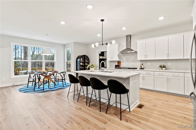 kitchen featuring white cabinetry, wall chimney exhaust hood, a kitchen island with sink, and hanging light fixtures