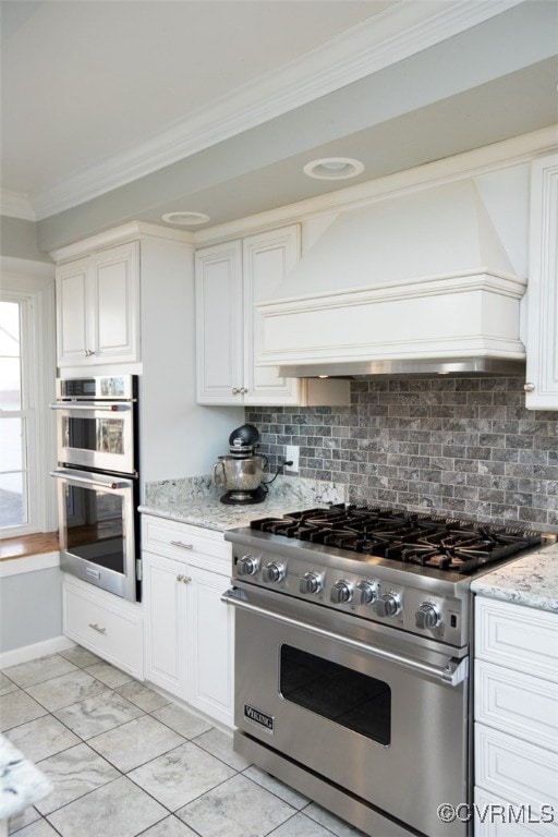 kitchen with white cabinets, decorative backsplash, appliances with stainless steel finishes, light stone counters, and custom range hood