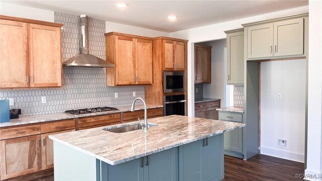 kitchen with appliances with stainless steel finishes, wall chimney range hood, sink, light stone counters, and a center island with sink