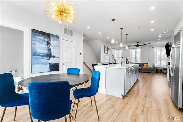 kitchen with hanging light fixtures, light wood-type flooring, stainless steel refrigerator, an island with sink, and ceiling fan with notable chandelier