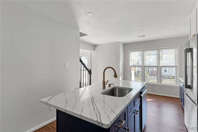 kitchen with light stone countertops, appliances with stainless steel finishes, blue cabinets, a kitchen island with sink, and sink