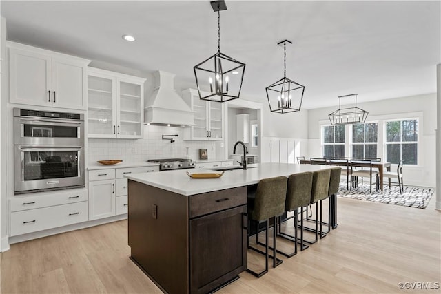 kitchen with appliances with stainless steel finishes, custom exhaust hood, a kitchen island with sink, white cabinets, and light hardwood / wood-style floors