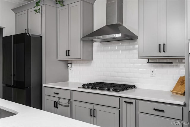 kitchen with gray cabinetry, backsplash, black refrigerator, wall chimney exhaust hood, and stainless steel gas cooktop