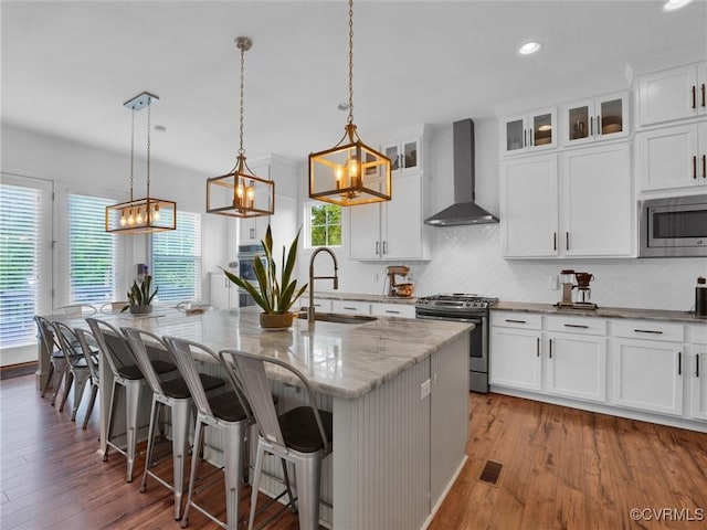 kitchen featuring pendant lighting, wall chimney range hood, light wood-type flooring, an island with sink, and stainless steel appliances