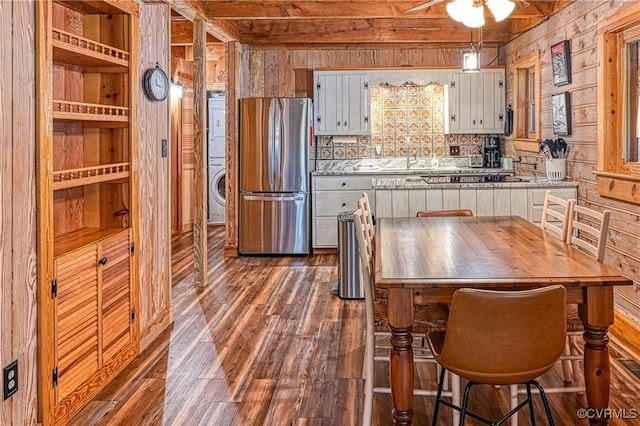 kitchen with wood walls, stacked washer and clothes dryer, dark hardwood / wood-style floors, beam ceiling, and stainless steel refrigerator