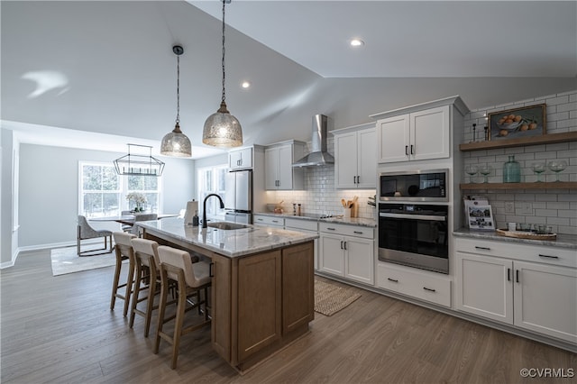 kitchen with wall chimney exhaust hood, an island with sink, pendant lighting, white cabinets, and appliances with stainless steel finishes