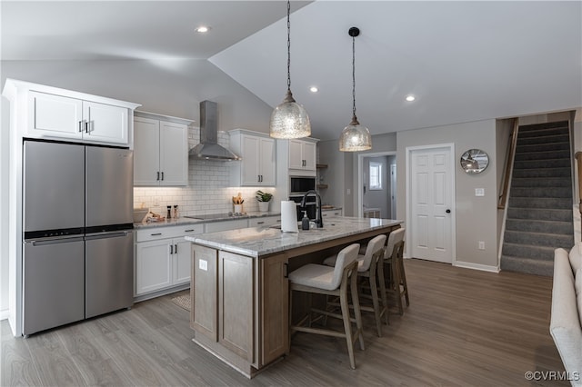 kitchen featuring a center island with sink, white cabinetry, stainless steel refrigerator, and wall chimney range hood