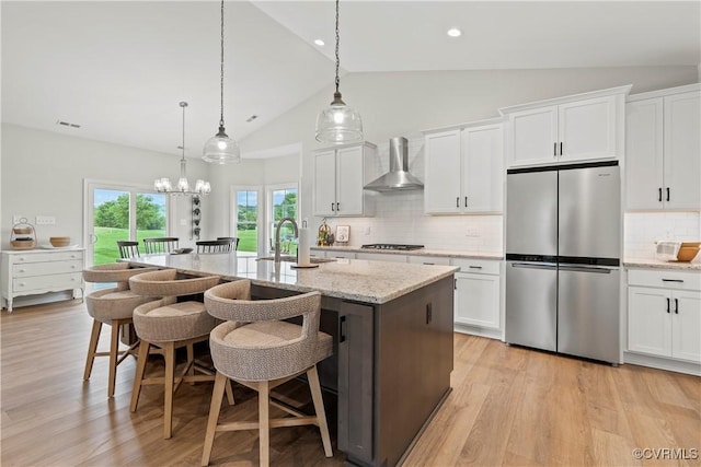 kitchen with appliances with stainless steel finishes, wall chimney exhaust hood, a center island with sink, decorative light fixtures, and white cabinets