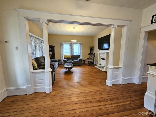 living room with wood-type flooring, a fireplace with flush hearth, baseboards, and ornate columns