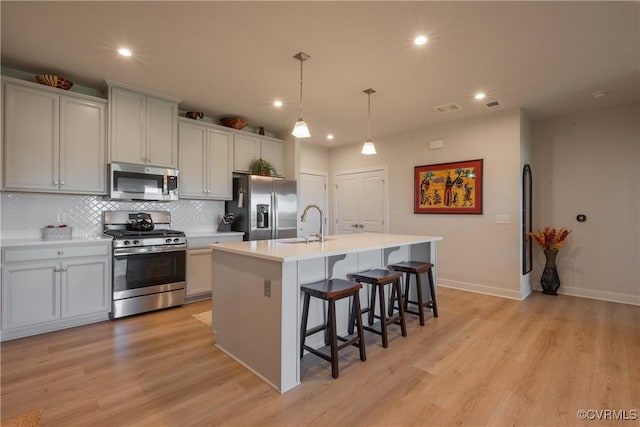 kitchen with decorative backsplash, an island with sink, appliances with stainless steel finishes, decorative light fixtures, and light hardwood / wood-style floors