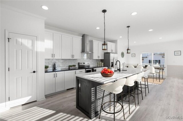 kitchen featuring pendant lighting, white cabinets, wall chimney exhaust hood, an island with sink, and stainless steel appliances