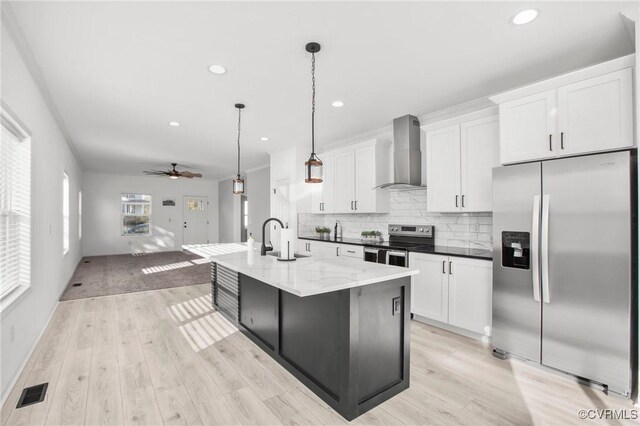 kitchen featuring white cabinets, wall chimney range hood, an island with sink, decorative light fixtures, and stainless steel appliances